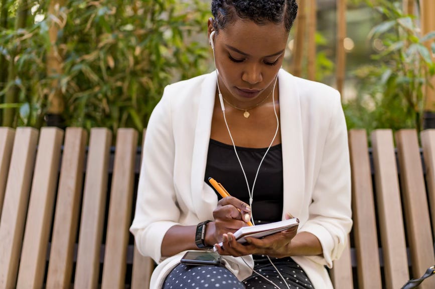 Black woman with earbuds in, sitting on a ebnch and writing on a notepad