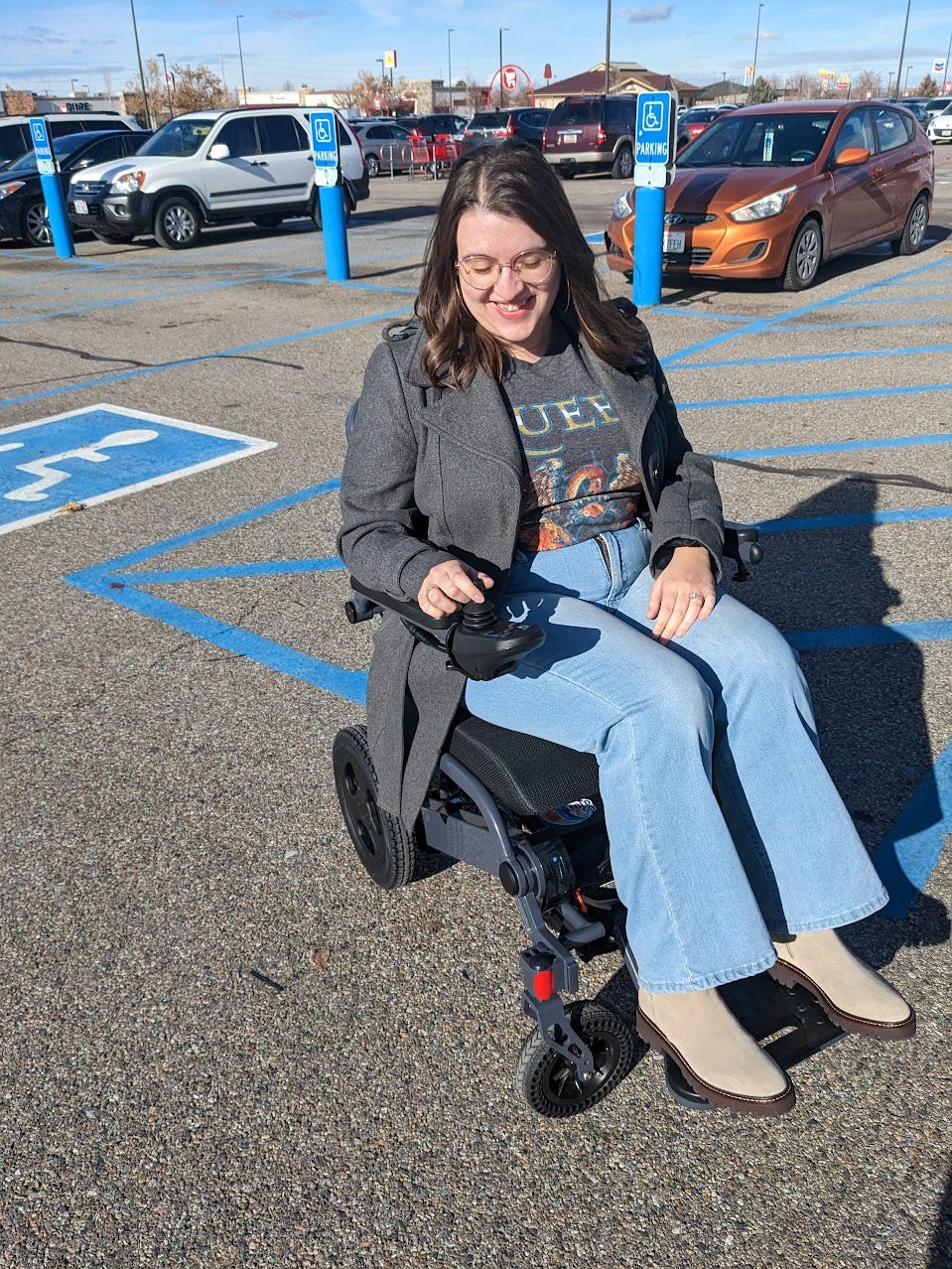Tessa, a white woman with brown hair wearing jeans, a Queen band tee, and a gray jacket, smiles while sitting in an electric wheelchair. She is in a parking lot, accessible parking spaces are visible in the background.