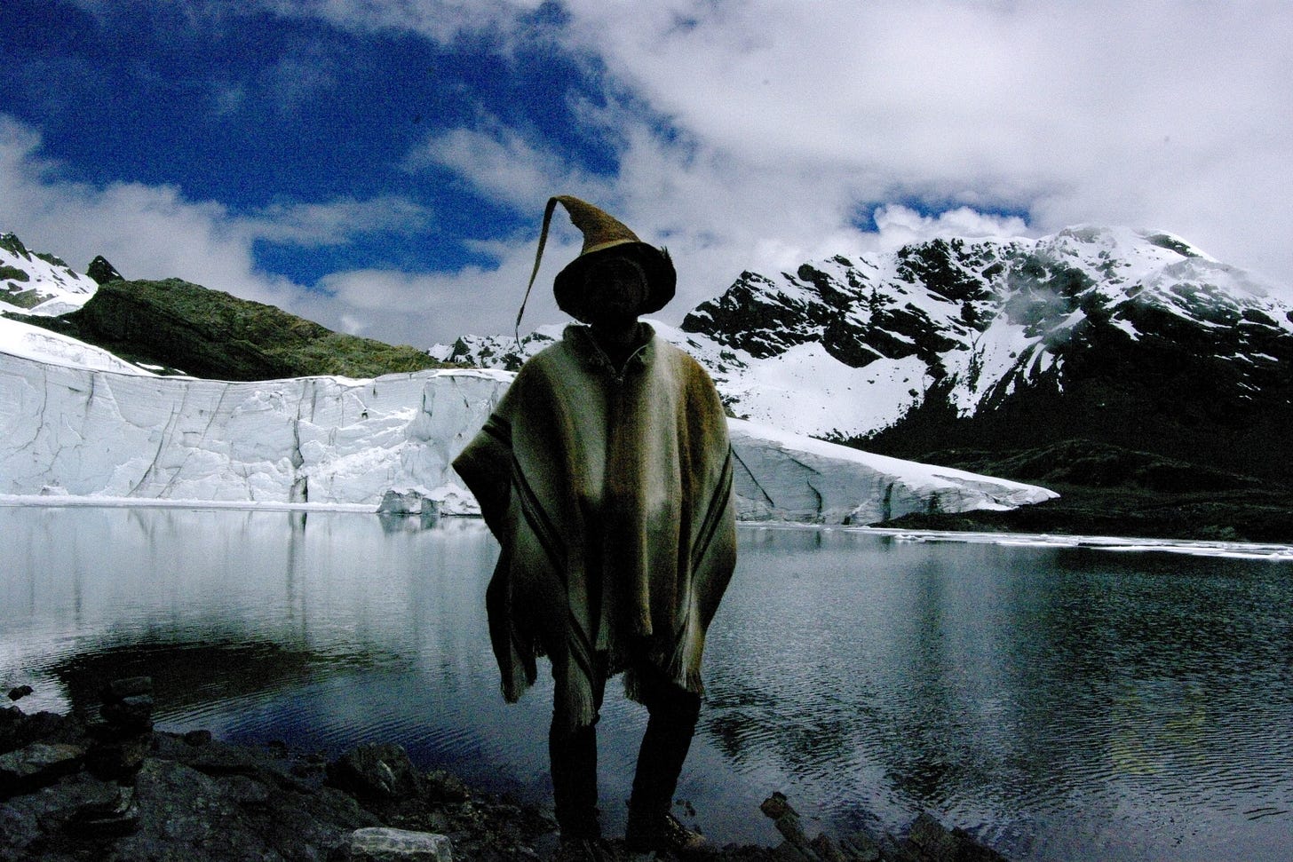 Eddie wearing a poncho and coconut skin hat with the silhouette of a wizard. He stands before glaciers and an alpine lake in the Northern mountains of Peru.