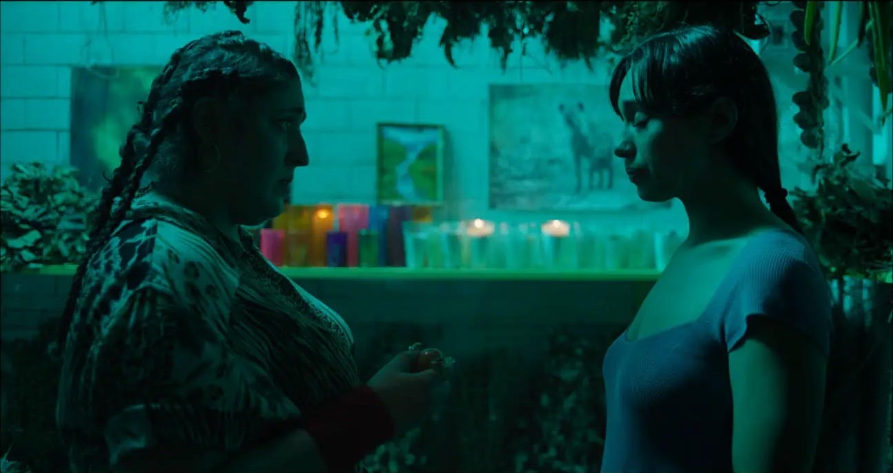 In a teal green lit room full of plants, two women face each other. The woman on the left, wearing numerous braids in her long brown hair, is holding something in her hands as she faces the young woman on the right, who is standing with her eyes closed.