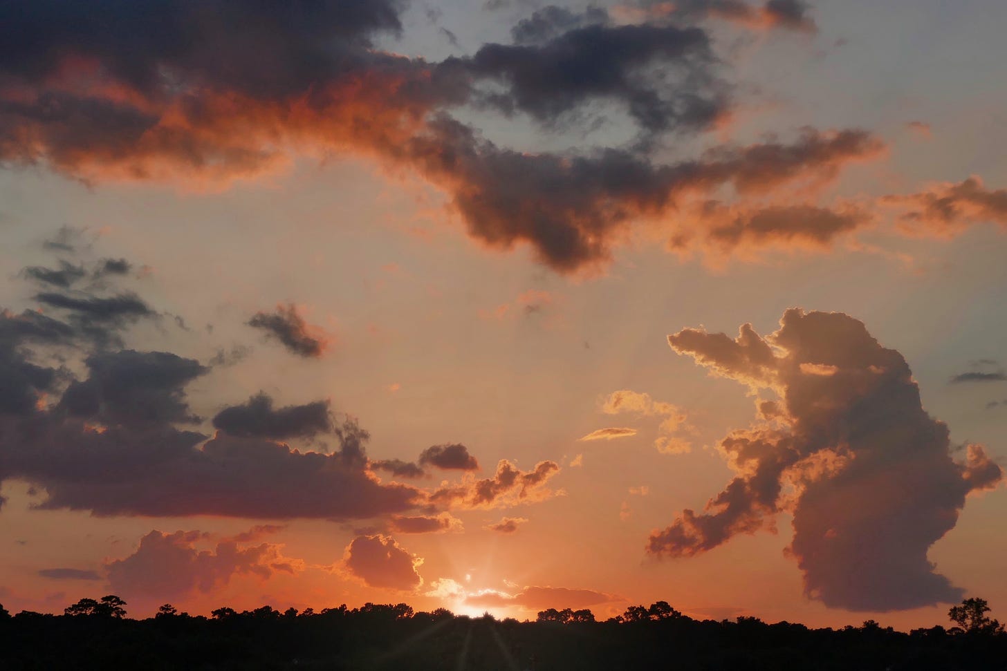 A sunset with the rays of the sun peaking above the horizon and the clouds and sky streaked with orange, corals, and grey