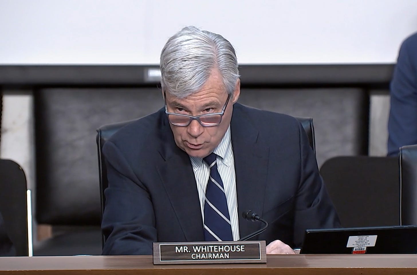 Wearing a dark blue suit, a US Senator with silver hair and glasses speaks at a desk. Before him is a plaque on which is written "Mr. Whitehouse, Chairman"