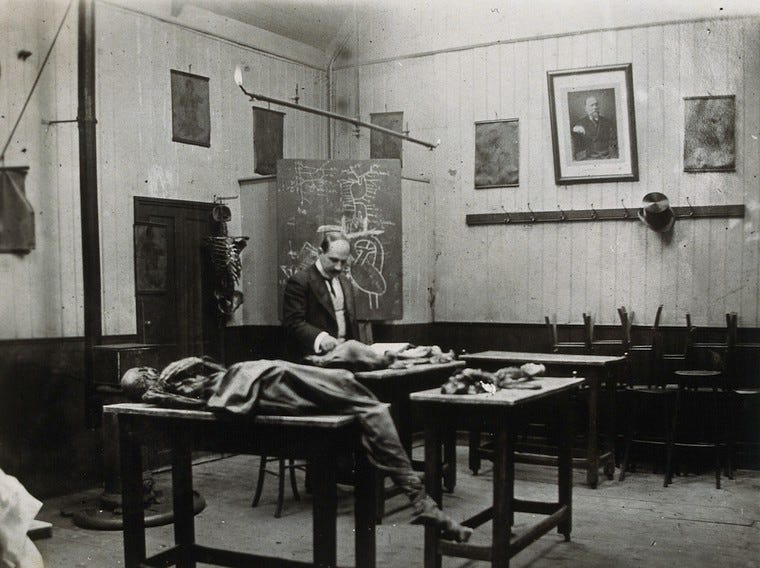 A black and white photograph of a bleak, cold-looking room with white tongue-and-groove panelling. There is a blackboard with anatomical diagrams. There are four small tables, on the nearest of which is a cadaver. A man in Edwardian clothing stands looking down at some body parts on another of the tables.