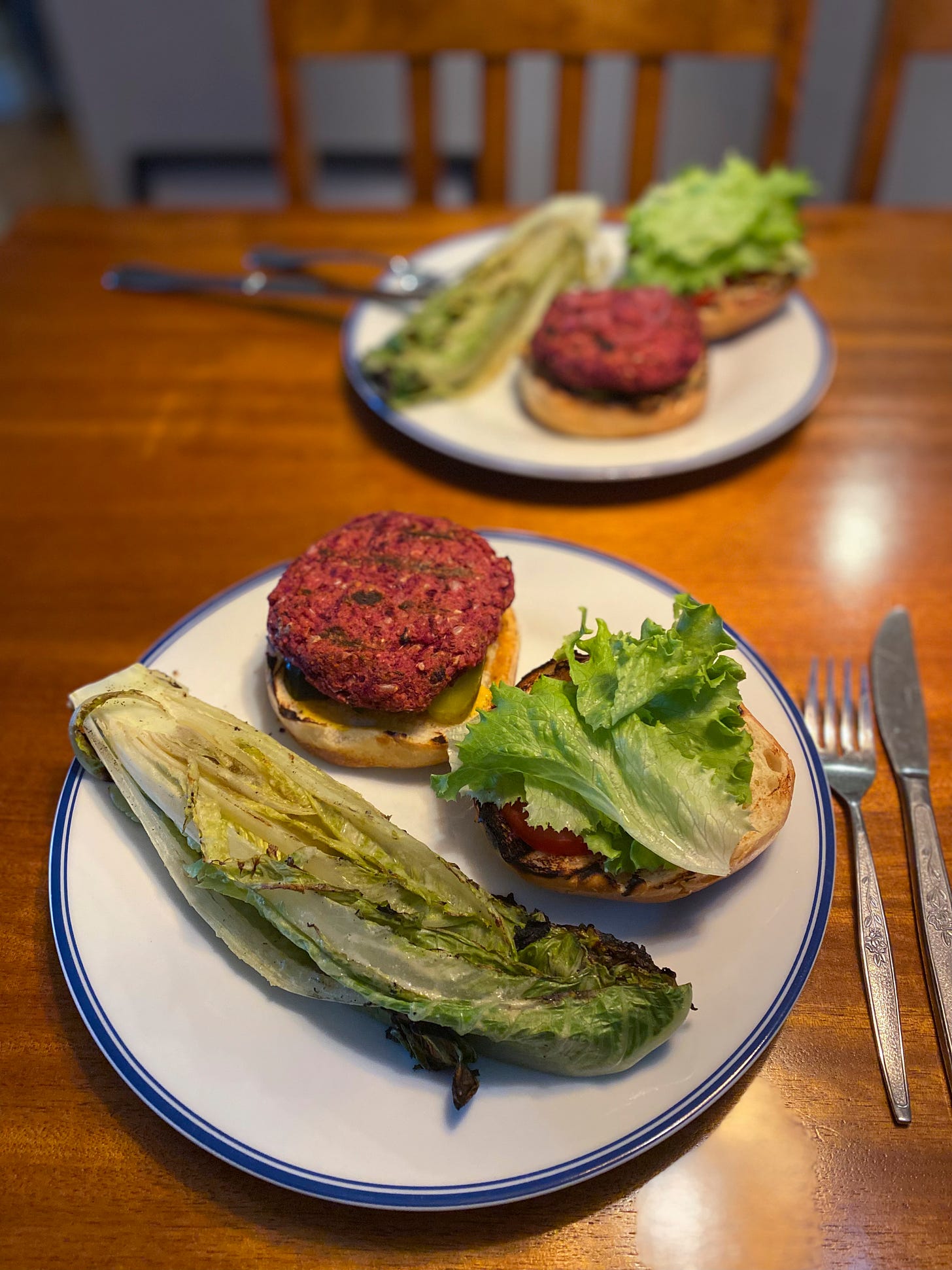 Two dinner plates, each with a burger on an open bun: one half holds the patty and pickle slices, and the other has tomato and lettuce. On the side is half a grilled romaine heart, in caesar dressing.