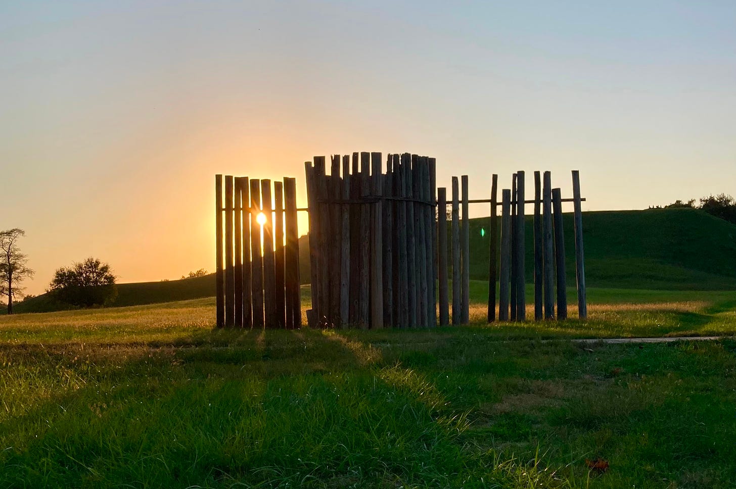 An orange sunset illuminates Monks Mound through a stand of wooden posts.