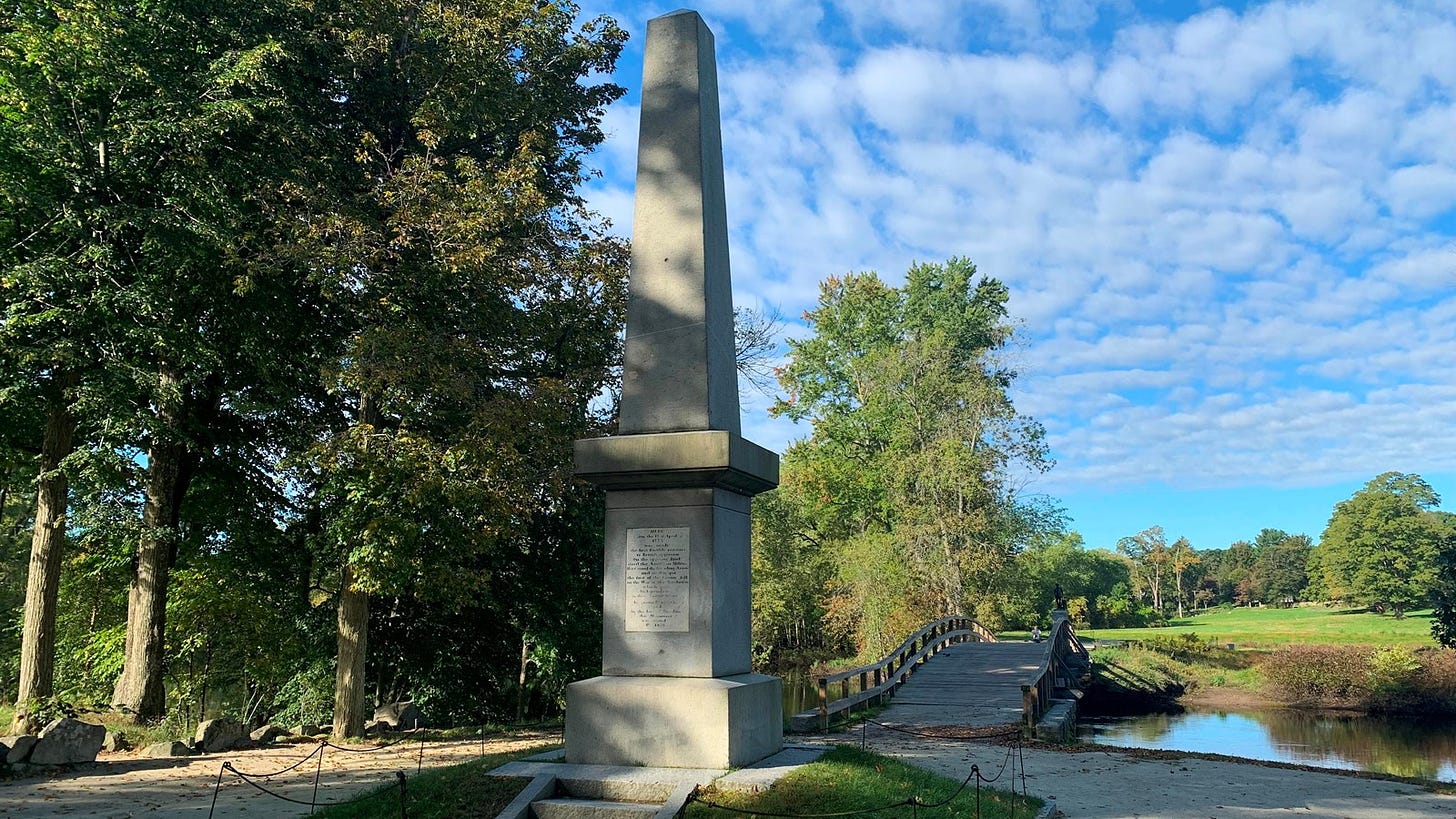 A stone obelisk monument 25 feet tall stands on a grassy mound in front of a wooden bridge.