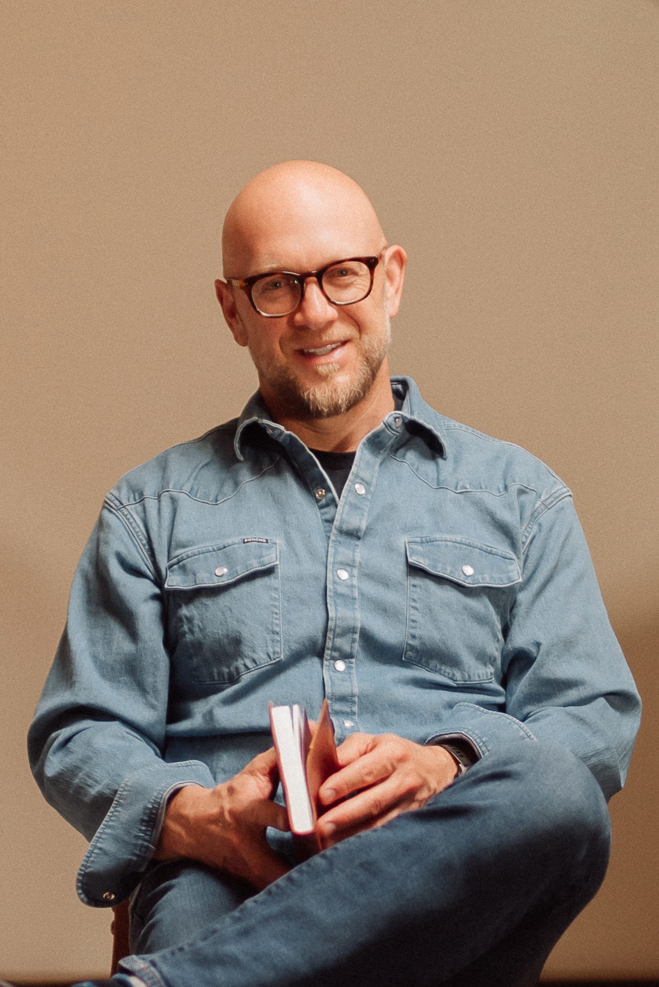 A bald man with a beard and glasses is sitting casually with his legs crossed. He is wearing a light blue denim shirt with buttoned pockets and jeans, holding a small, open book in his hands. He is smiling and looking directly at the camera, exuding a relaxed and approachable demeanor against a plain background.