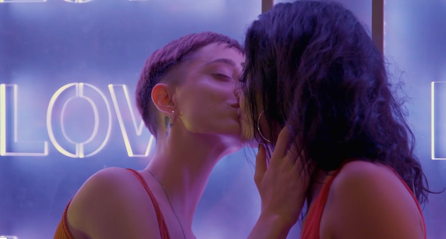 A slender white woman with short hair and a slender long neck leans forward as she kisses a woman with wavy brown hair as she touches her neck in front of a softly lit lavender-hued background.
