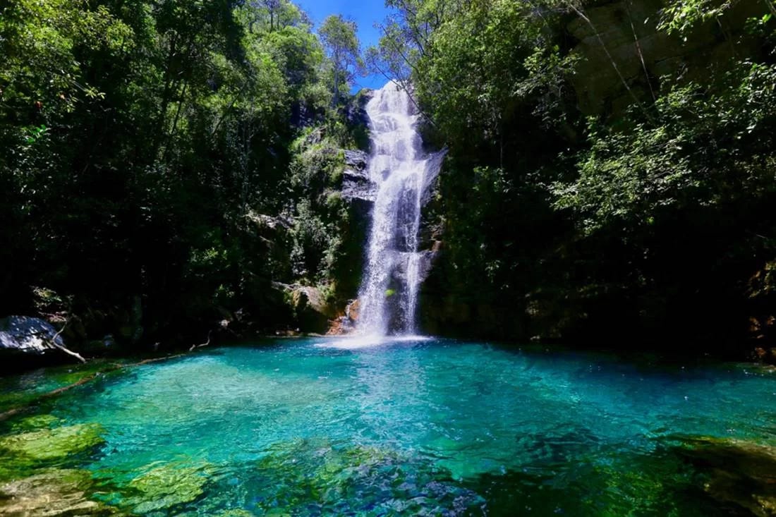Uma cachoeira desce em meio à vegetação, desaguando num lago azul cristalino. A foto é muito bonita, com tons azuis e verdes dos mais variados.