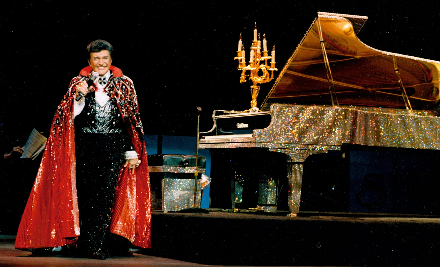 Next to a completely gold crystal bedazzled piano, on which is an elaborate candelabrum, stands Liberace, a white man wearing a sparkly black suit with an elaborately bejeweled red cape.
