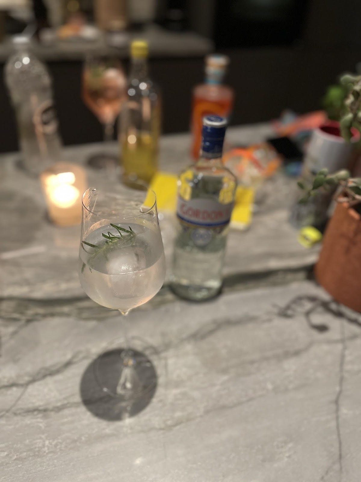 Photo of a marble countertop with a glass of gin & tonic in the foreground. Behind it are blurred bottles and a candle.