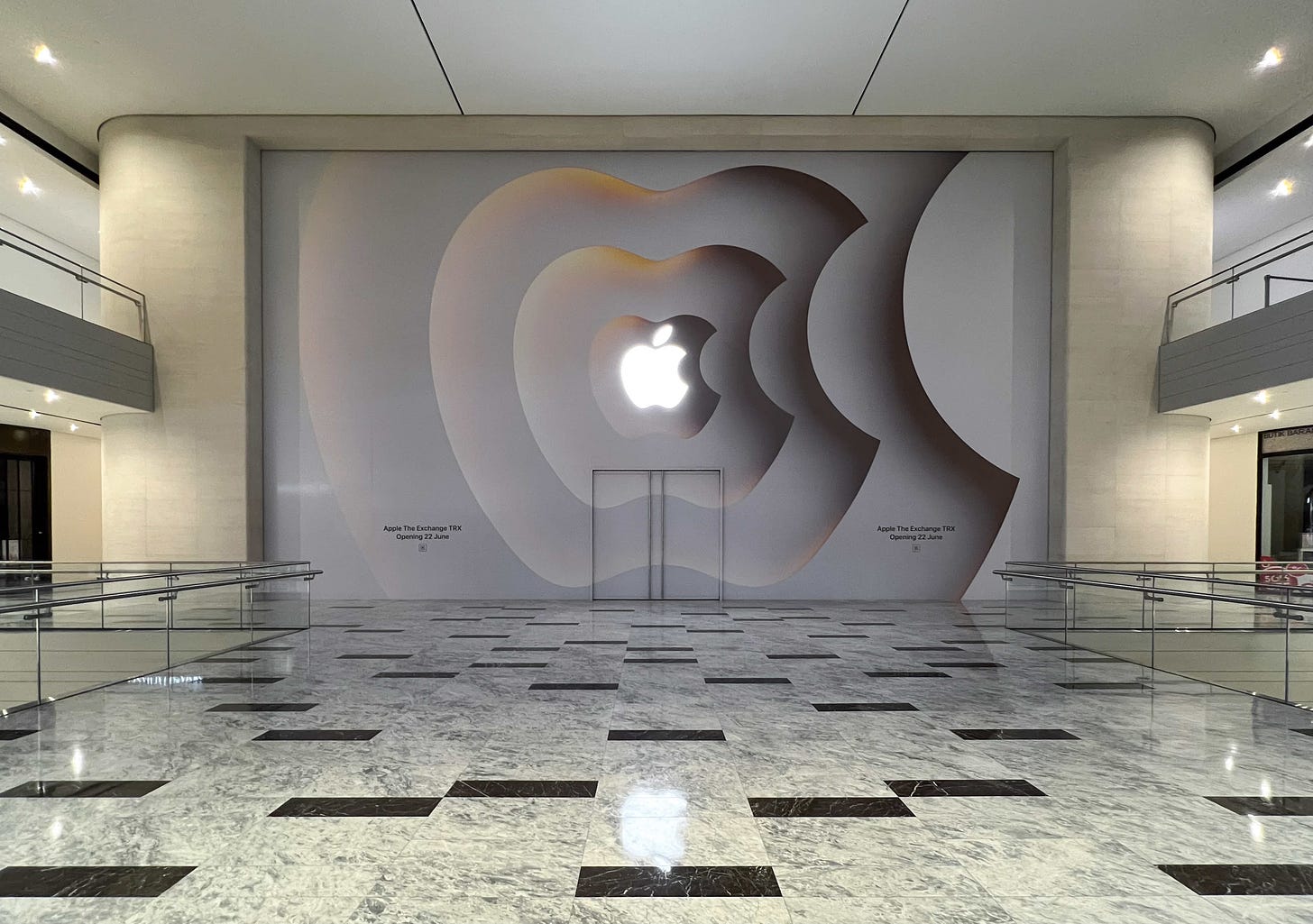 The indoor entrance to Apple The Exchange TRX spans two floors inside the mall. A glowing Apple logo is centered on the glass between the two levels.