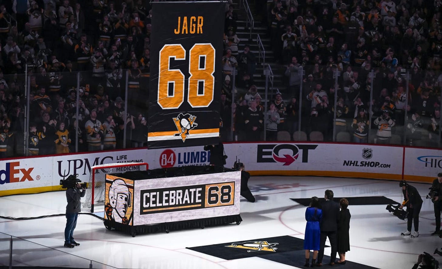 Jaromir Jagr and his family stand on black carpet on the ice while watching a large banner with the word Jagr and the number 68 on it as it is raised out from a box that says Celebrate 68