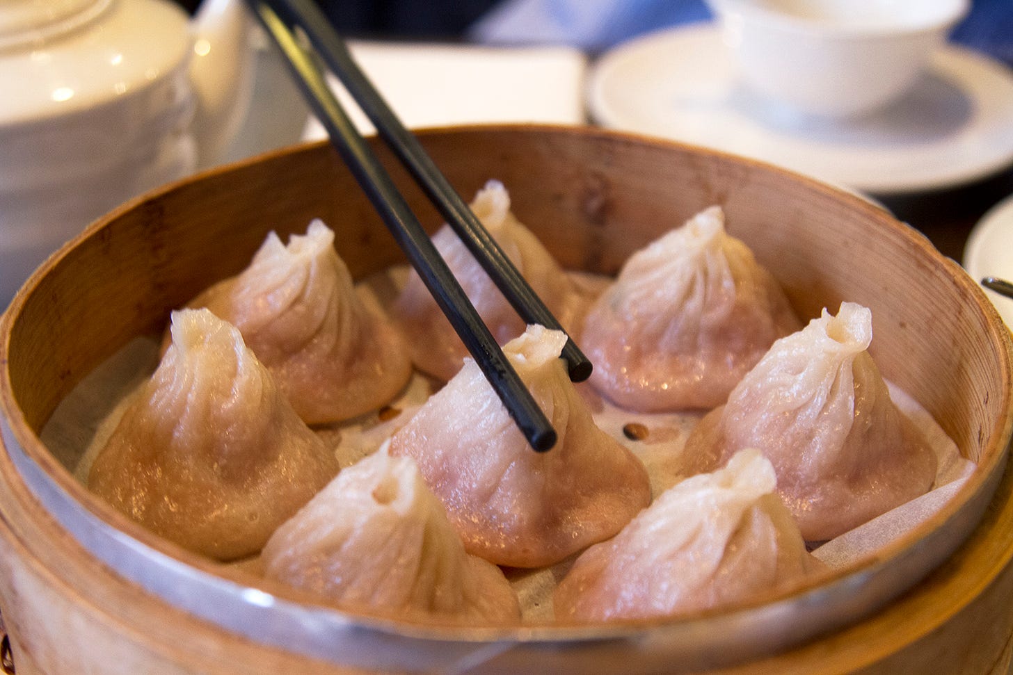 Eight dumplings sit in a bamboo steamer, with a pair of chopsticks reaching for one of them.