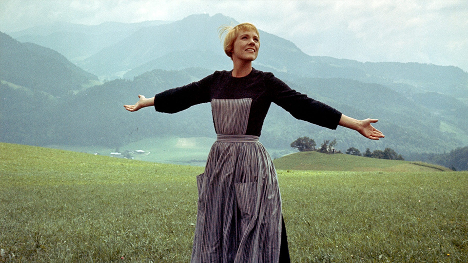 Julie Andrews como Maria, com uma blusa de mangas compridas prestas e um avental acinzentado com bolsos. Ela é loira e tem cabelos curtos e está com os braços abertos olhando para cima e para sua esquerda. Ela está em um campo verde numa região montanhosa.