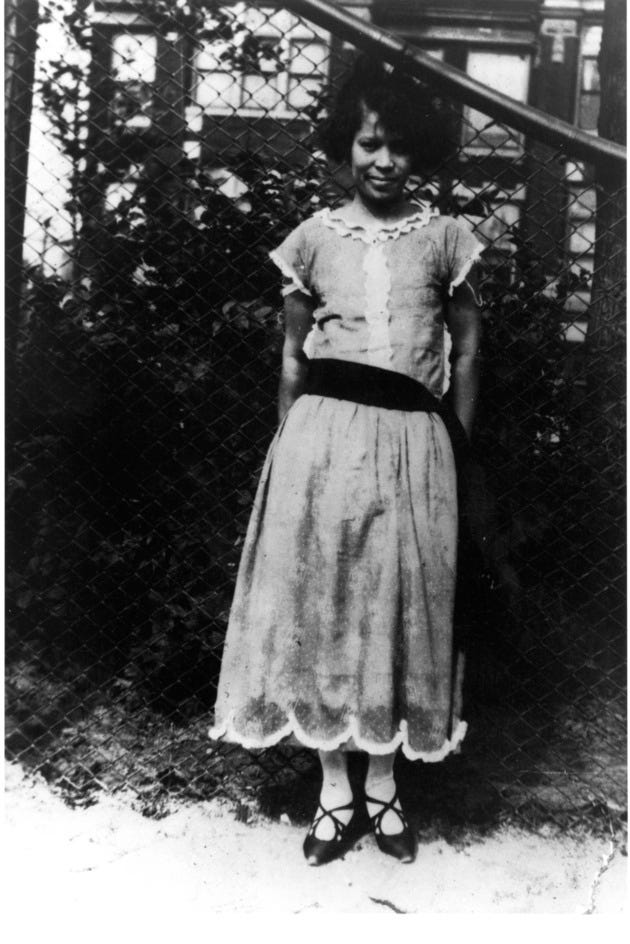 YOUNG ZORA NEALE HURSTON, OUTDOORS. LEANING AGAINST A CHAIN-LINKED FENCE, WEARING A LIGHT DRESS WITH BLACK SASH AT THE WASTE. SMILING AT CAMERA.