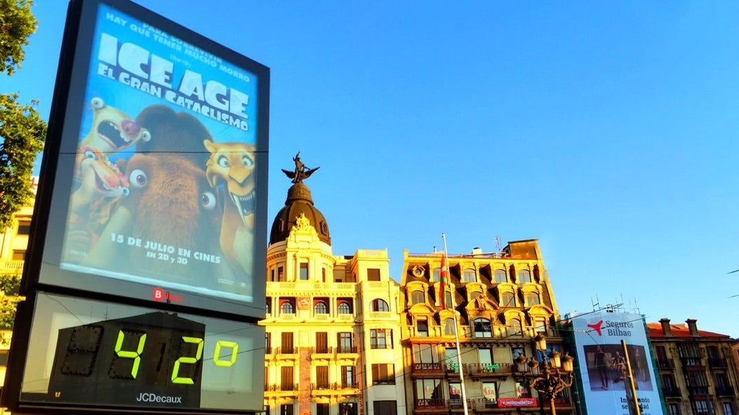blue sky, old Spanish buildings in the background. On the left is a movie poster announcing Ice age, and right below is a sign indicating it is 42 celsius
