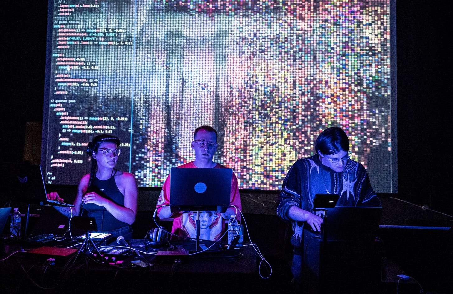 3 DJs on their laptops in a dark club, with code on a screen in the background