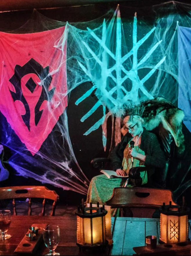 Elle in costume with a book in her hand and microphone in the other sitting on a large leather chair with a fur draped over it; behind her, the wall has a Horde banner and nordic symbol painted on a black wall with webs stretched out over everything; there is also a greenish hue because of the lights