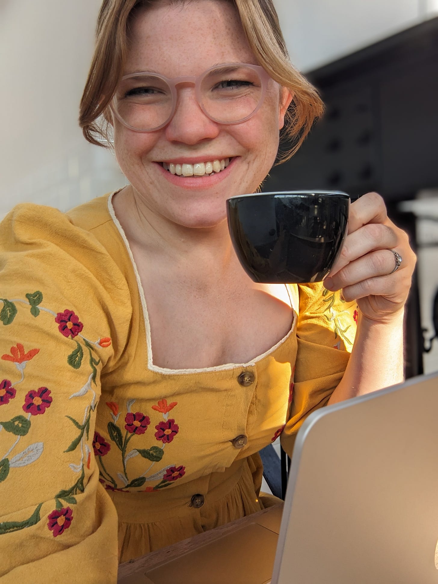 Libby Page sat at a desk with her laptop and a cup of tea, dressed in a yellow dress covered in embroidered flowers. She is smiling and wearing glasses.