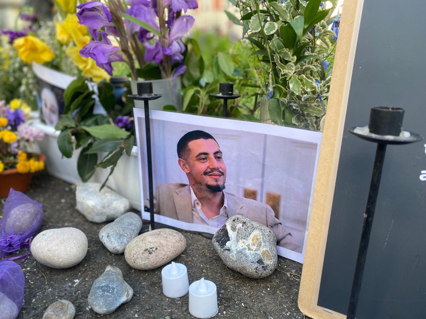 A4 photograph of a young man with a beard and dark hair in a beige suit laughing. The photo sits in amongst flowers and stones and candles at a memorial for the victims of the Hamas attacks on 7 October in Israel. The memorial is located in Brighton & Hove in the UK.