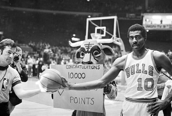 A man wearing a basketball uniform receives a ball from another man on a court. A mascot stands behind them, holding a “Congratulations” sign.