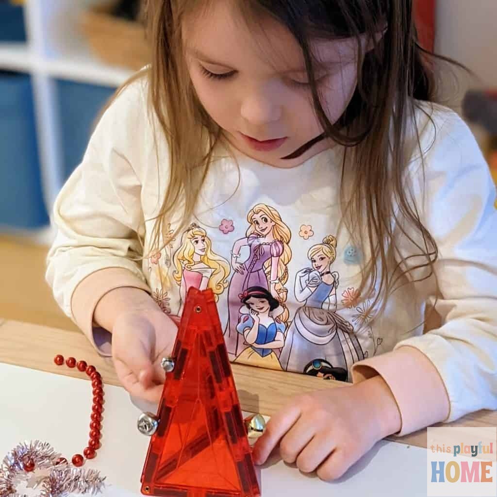 a 5 year old girl has made a red christmas tree from red magnetic triangles, she is adding jingle bells to decorate the tree 