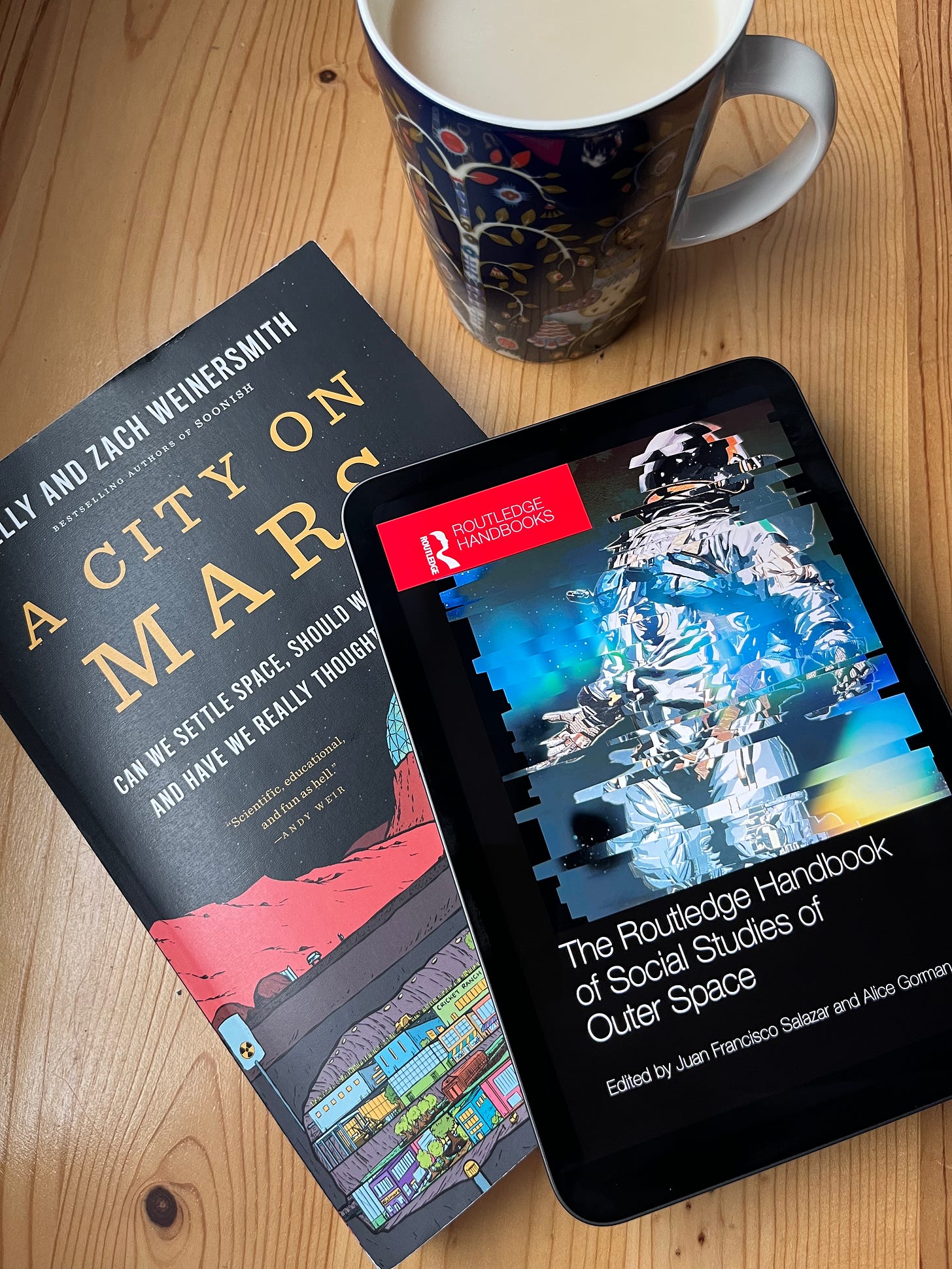 An iPad displaying the cover of the Routledge Handbook sits on top of a paperback copy of A City on Mars, both resting on a wooden surface next to a blue mug of tea.