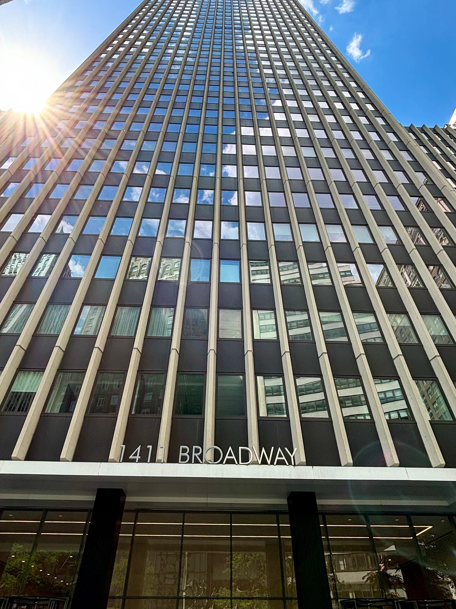 Looking up at 1411 Broadway. It is a large glass 60s style office tower reflecting the blue sky and clouds. The sun's rays create lines of force from behind the left side of the building.