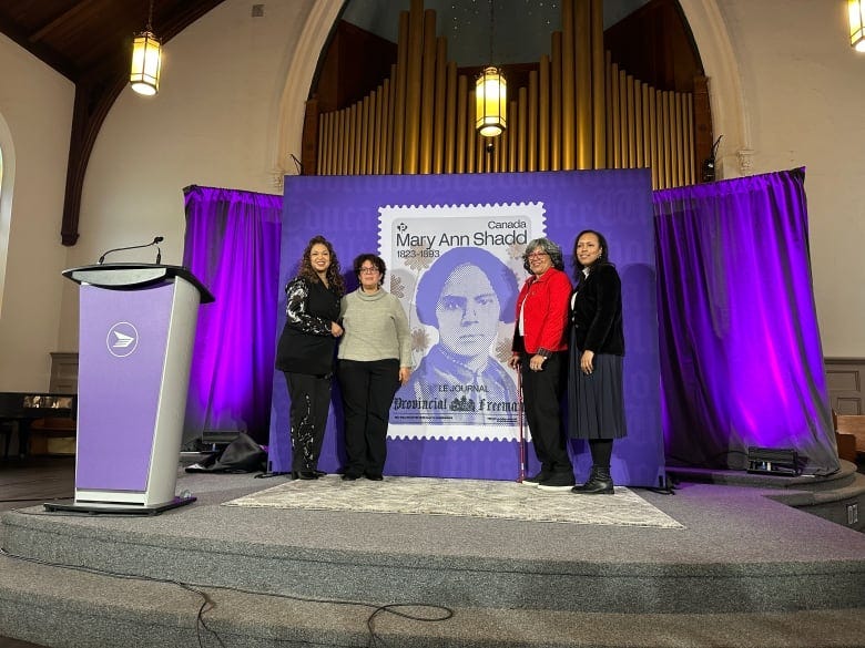 Four women stand next to a large image of a stamp