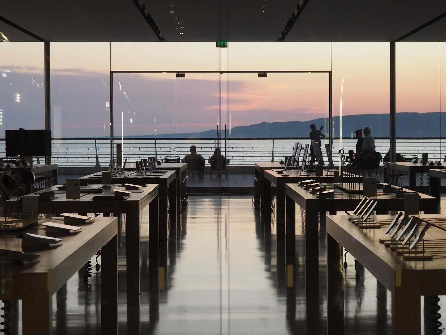 A moody scene at Apple Marseille. The sun is setting, the store is dim, and people sit near the water taking in the beauty of nature.