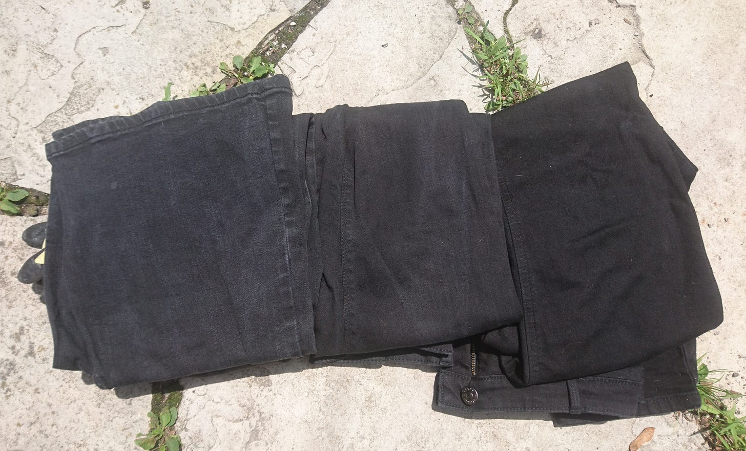 three pairs of black jeans folded into quarters and lined up on flagstones with moss between the cracks