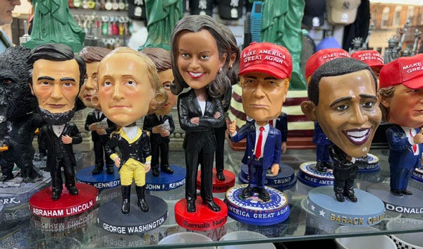 A shelf of bobbleheads in a souvenir shop. Abraham Lincoln and George Washington are on the left. Kamala Harris is in the center, taller than the rest. On the right are several Trump figures with red hats and an Obama with a larger head than any other.