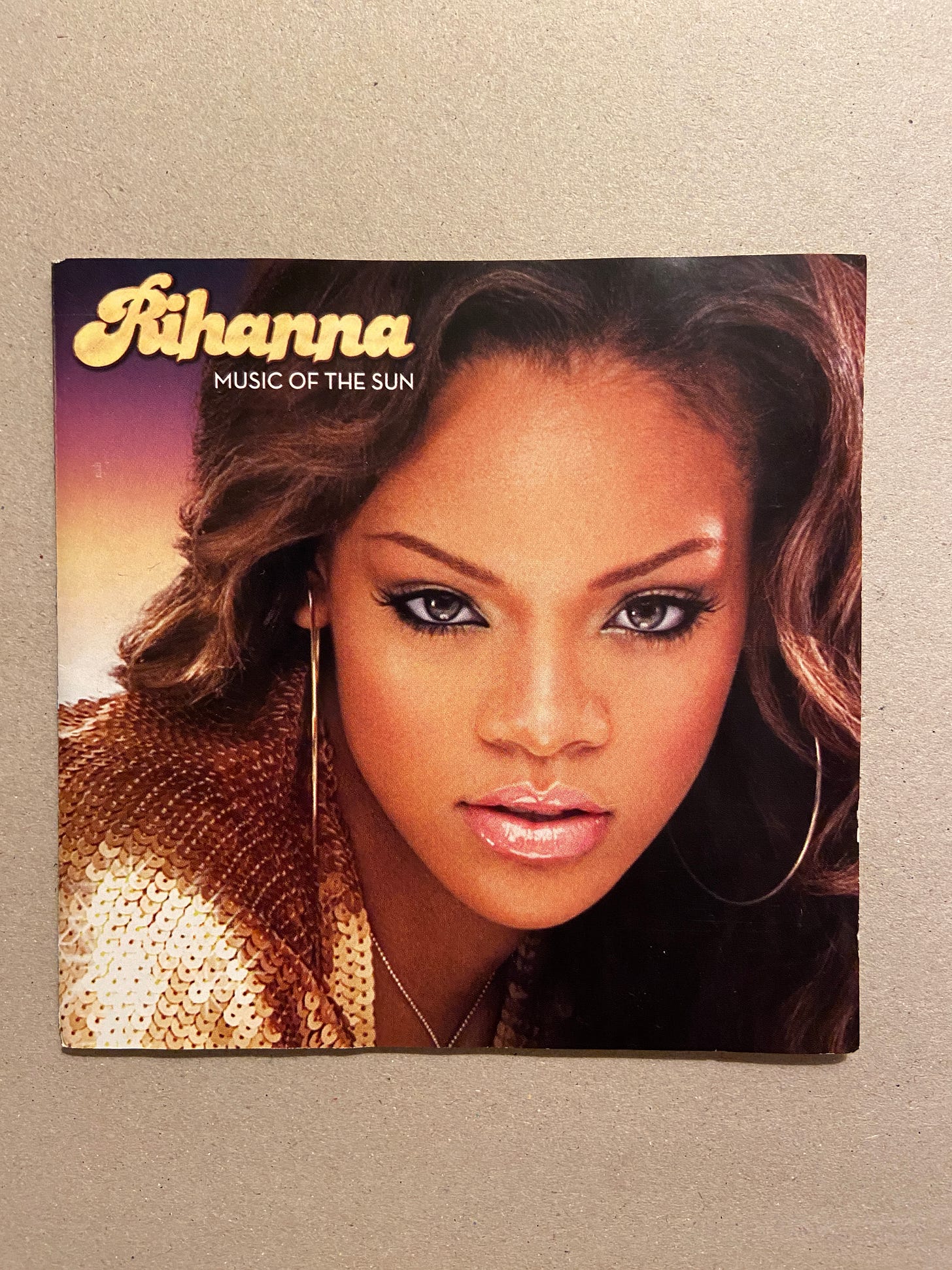 the cover for Rihanna's debut album, Music of the Sun. she stares into the camera while wearing gold hoop earrings and sequin top.