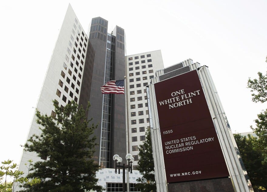 View looking up at a multistory tower building, built in a modern style, with what appears to be concrete and marble or other stone. A sign with the agency's name is in the foreground.