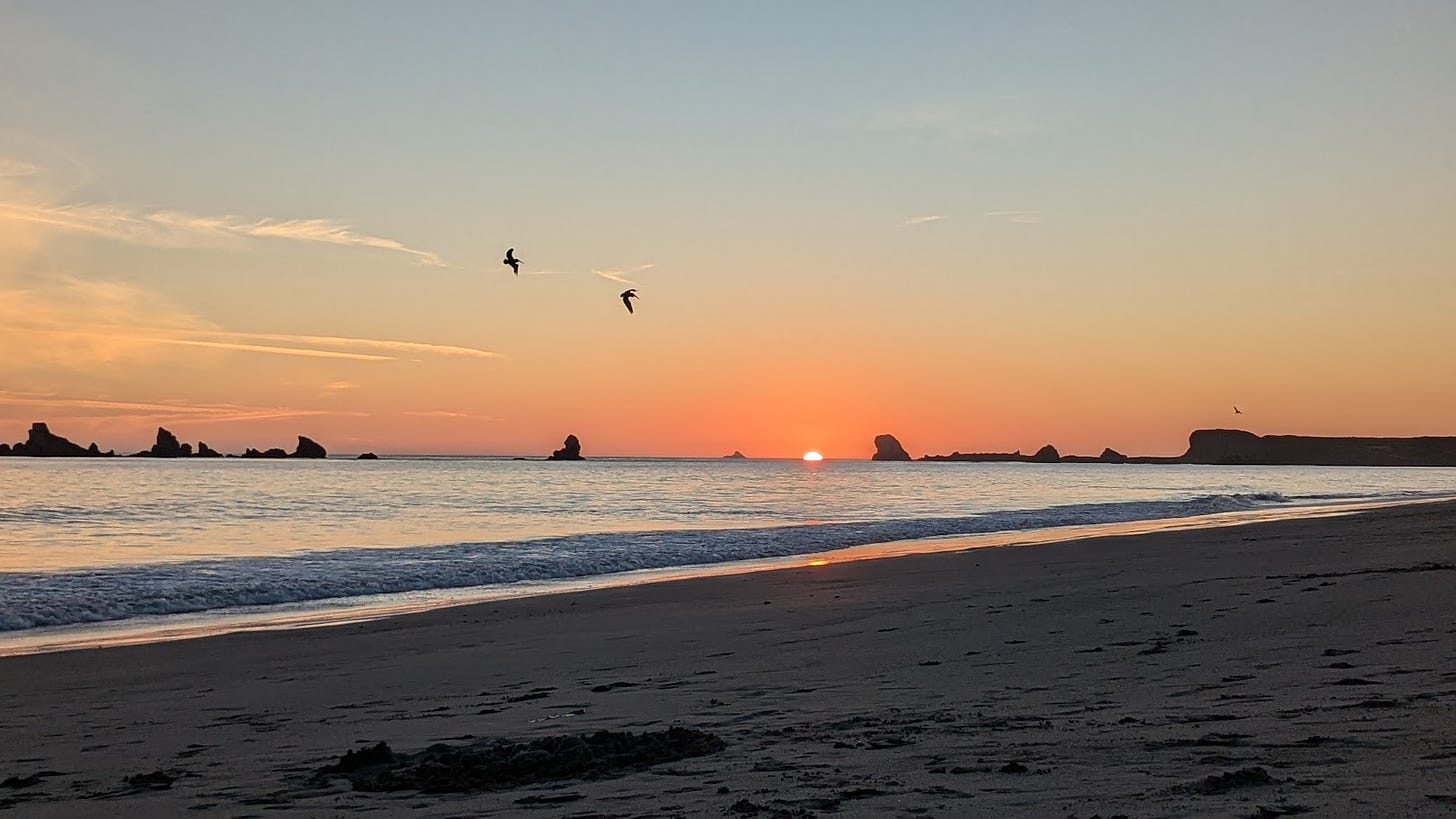 Red orange sunset behind rocky ocean outcroppings, ocean waves, and a sandy beach