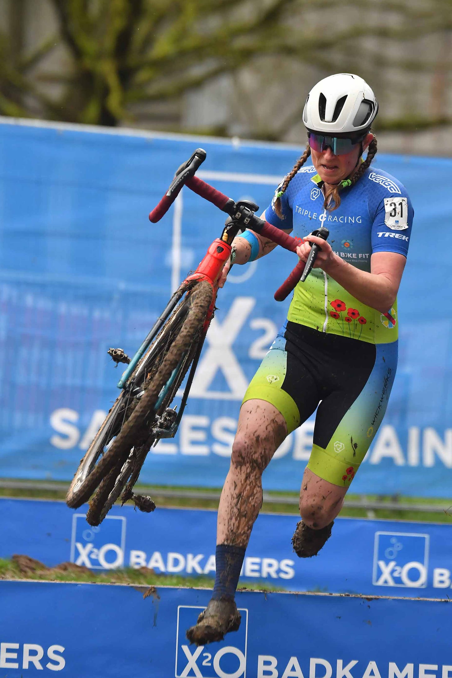 A female cyclist lifts her red bike while leaping over a barrier on a muddy cyclocross course.
