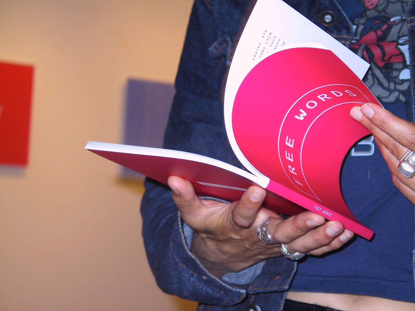 A closeup photograph of someone looking through a copy of a hot pink book with the title Free Words. The person is wearing a blue T-shirt under a jean jacket, and has rings on several fingers.