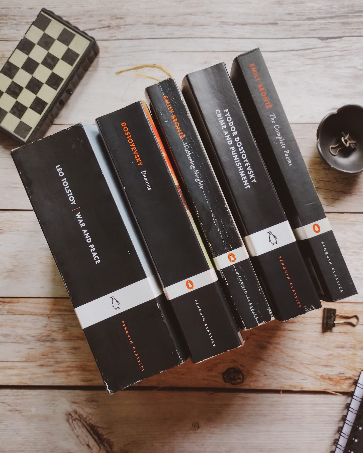 Five Penguin black classics aligned on a wooden table with their spines showing, War and Peace by Leo Tolstoy, Demons by Fyodor Dostoevsky, Wuthering Heights by Emily Brontë, Crime and Punishment by Fyodor Dostoevsky, The Complete Poems by Emily Brontë