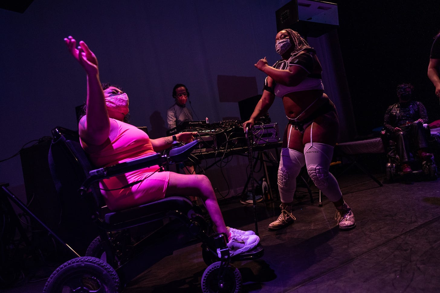 Two dancers in shorts and crop tops, one of whom is using a wheelchair, move to the music while a DJ spins. 