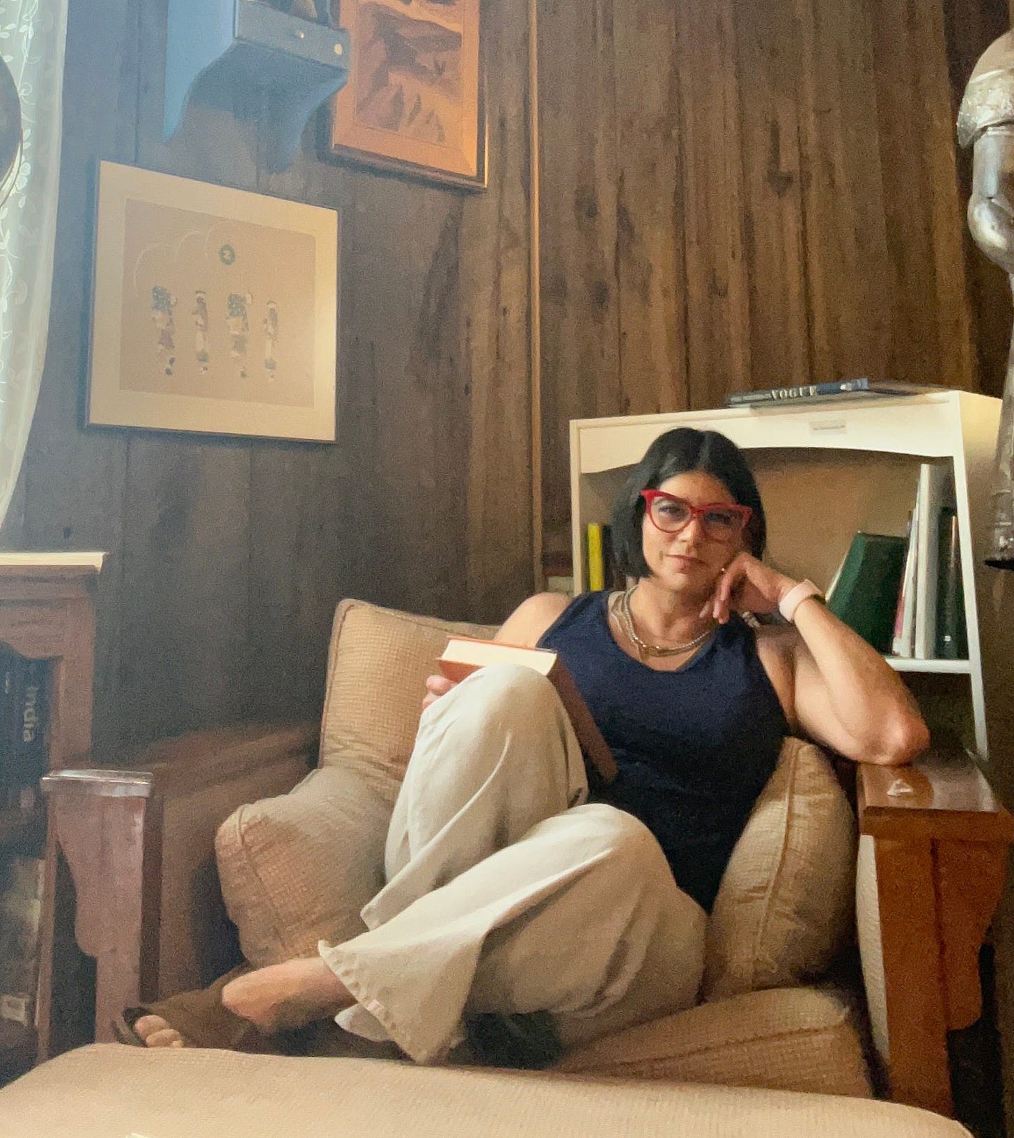 A dark-haired woman with red eyeglasses, sitting in a comfy chair with a book, looking at the camera, surrounded by other books and art on dark paneled walls