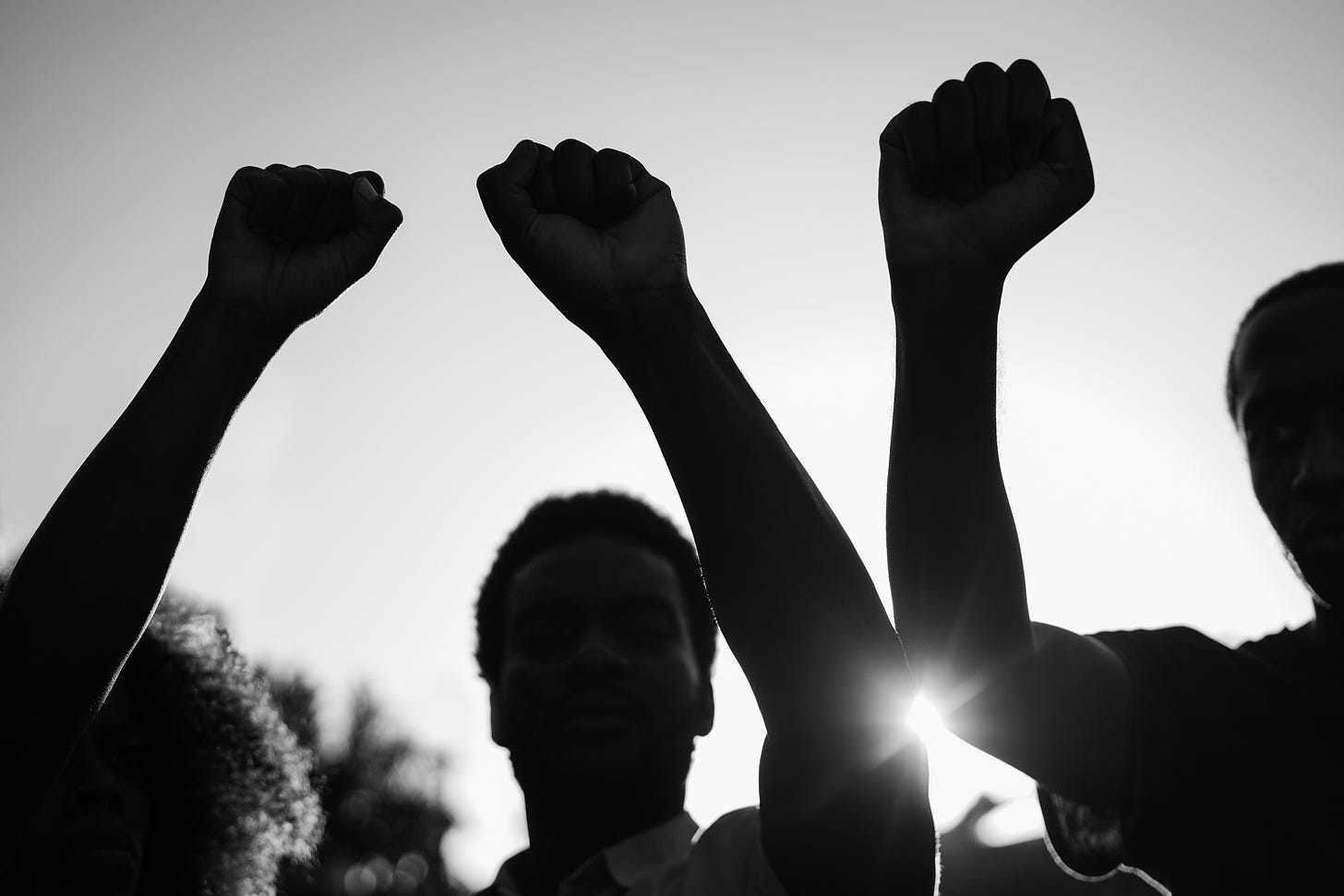 A black and white image of three protestors, in silhouette, with their fists raised high as the sun fades in the background. 