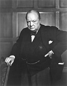 Churchill, 67, wearing a suit, standing and holding into the back of a chair