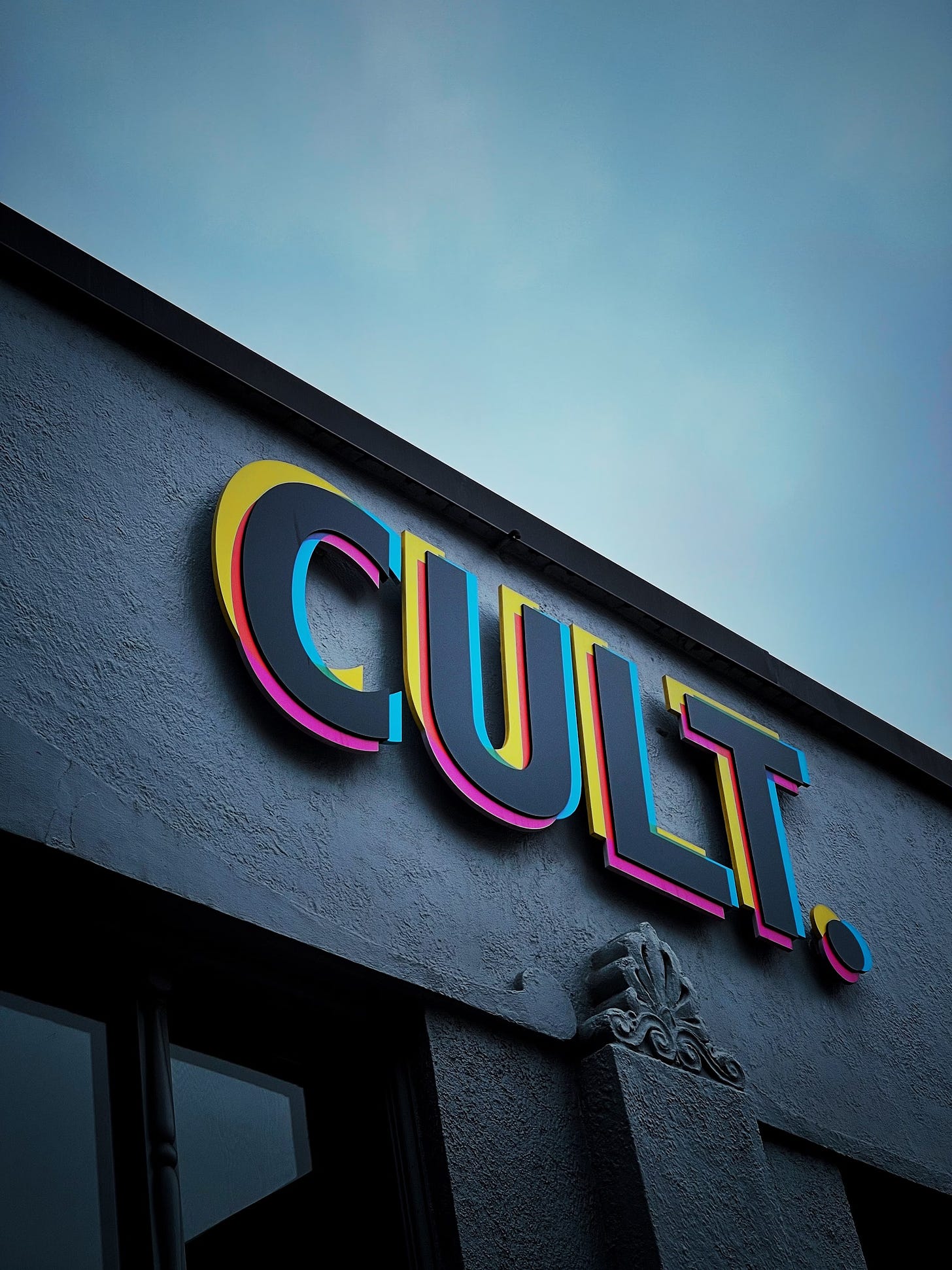 The word "CULT" in big black block letters (with yellow and pink showing underneath) on a stucco facade.