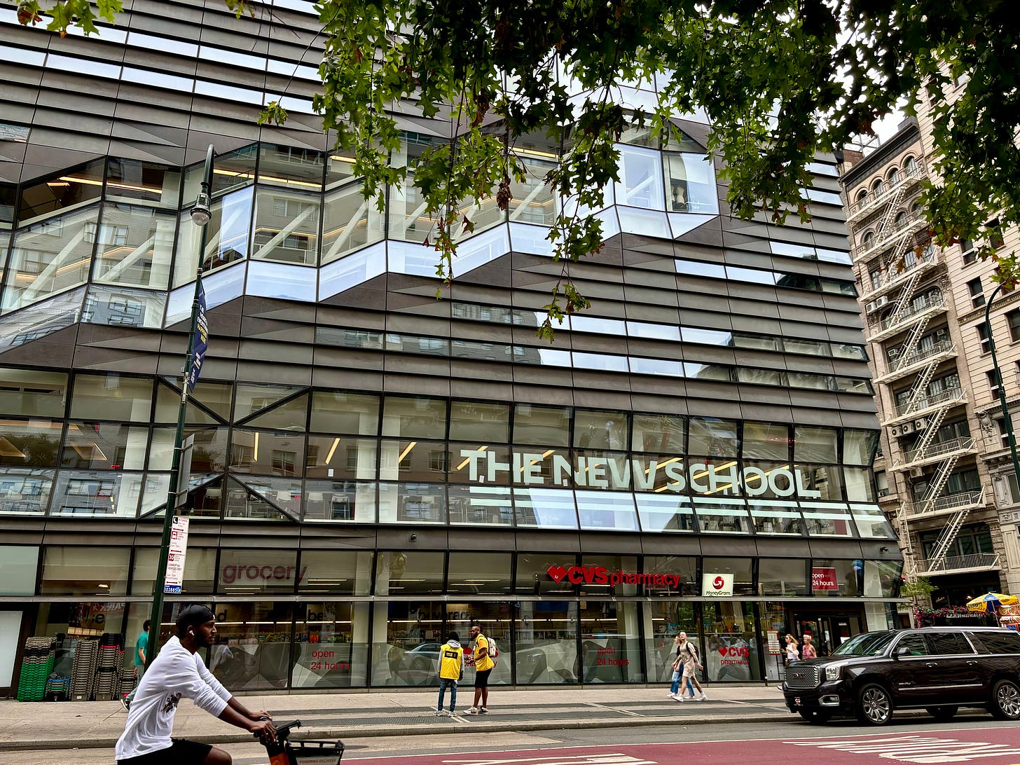 A glass and metal-facade building with angles jutting out and a diagonal row of windows. A tree full of leaves fills the upper, right-hand corner. The right is the wide street of 5th Avenue. A biker is zipping by.