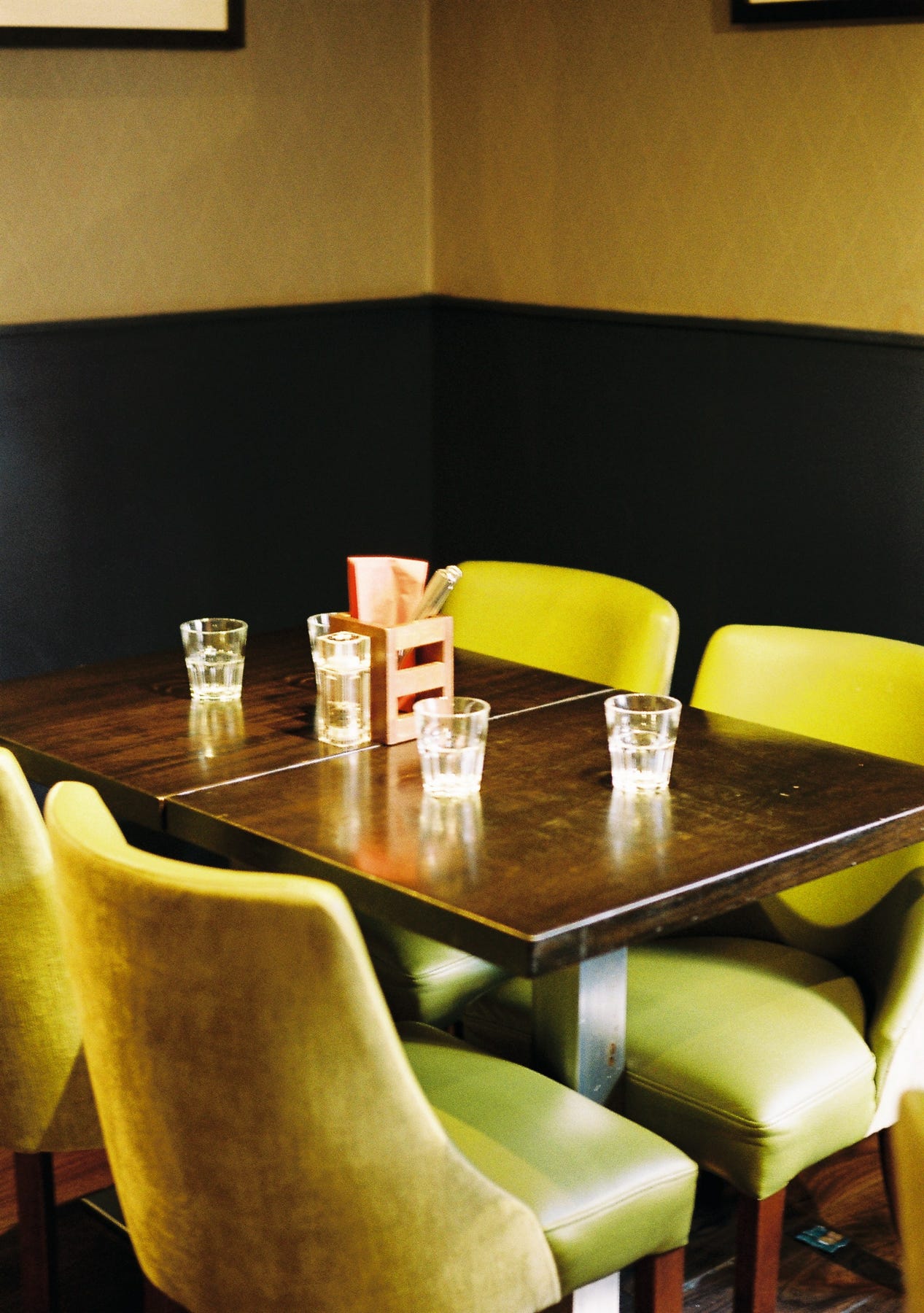 An empty table in a bar restaurant with water glasses. The chairs a greenish yellow, the wall dark at the bottom with dark yellow wallpaper above.