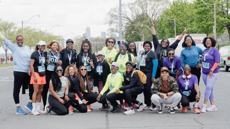 A group of 21 women in athletic clothes stand on a closed off downtown Toronto street, smiling for the camera.