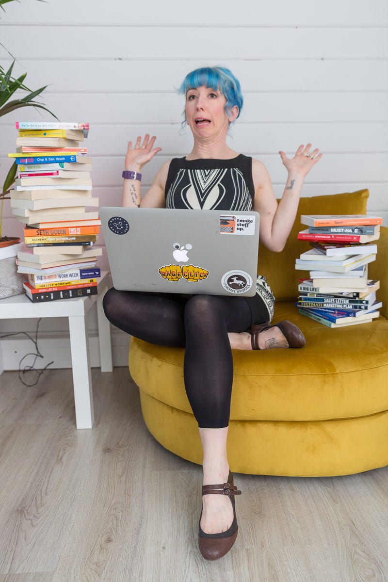 Vicky sits on a yellow comfy chair, surrounding by tottering piles of books, laptop on knee, hands up, and 13 chins as she leans away from her mess.