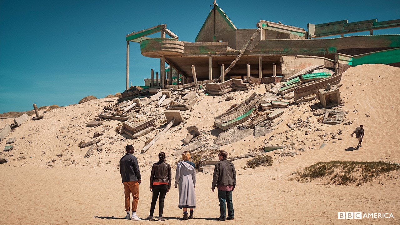 This image shows Team TARDIS (from left to right, Ryan Sinclair, Yaz Khan, the Thirteenth Doctor, and Graham O'Brien) standing outside a deteriorating building in the middle of the planet Desolation's sandy desert. Debris from the building litters the large sand dune in front of them. This image shows Team TARDIS (from left to right, Ryan Sinclair, Yaz Khan, the Thirteenth Doctor, and Graham O'Brien) standing outside a deteriorating building in the middle of the planet Desolation's sandy desert. Debris from the building litters the large sand dune in front of them.