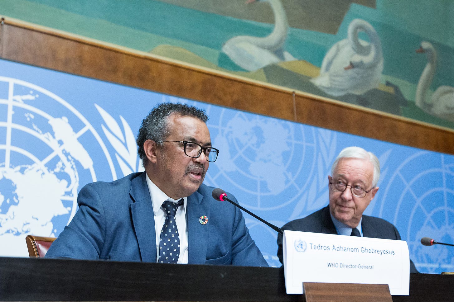 The head of the World Health Organization Dr. Tedros Adhanom speaking into a lit microphone at the UN office in front of a blue and white banner with the WHO logo in a repeating pattern